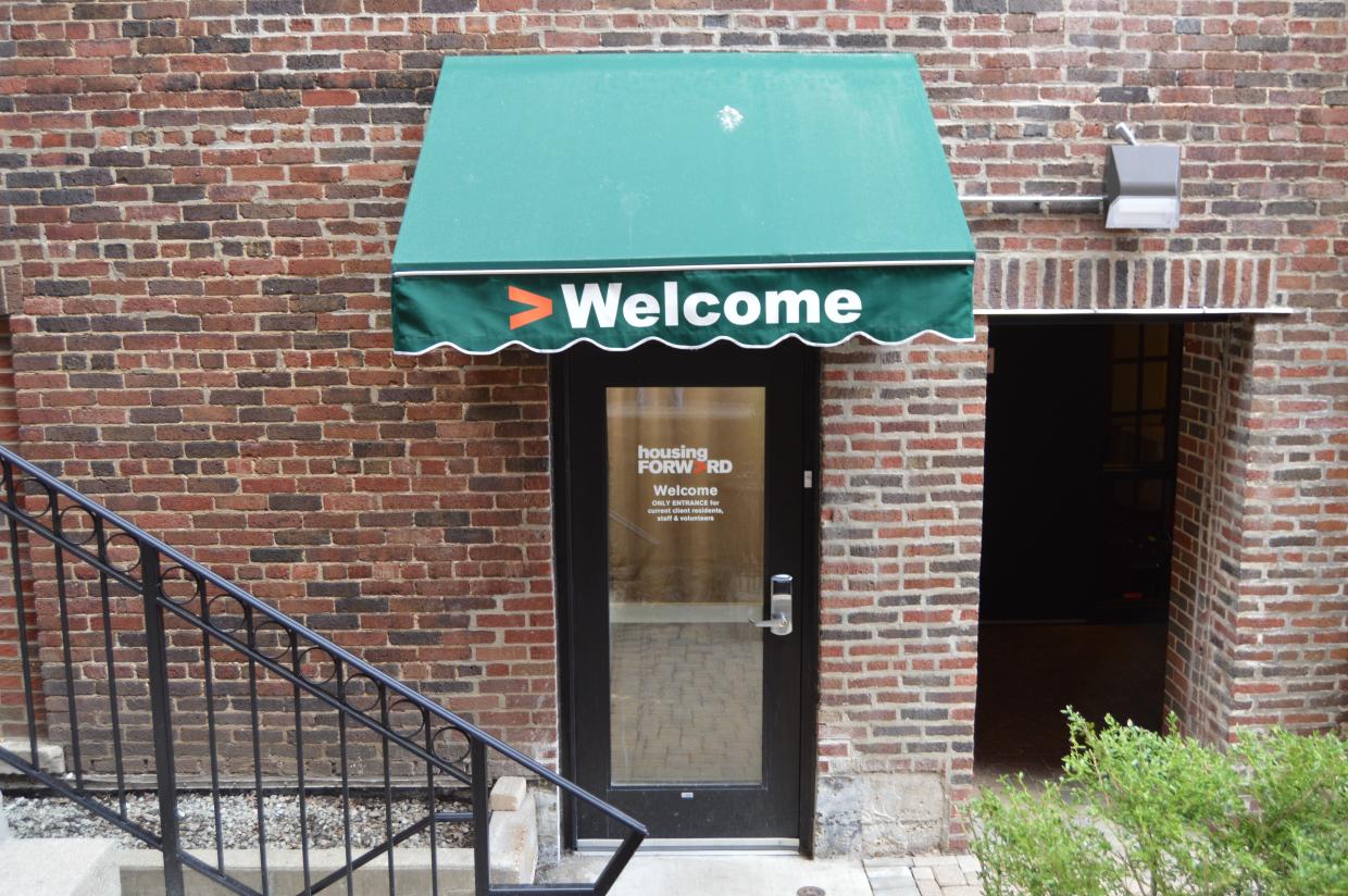 Color photograph of the façade of a brick building with a door. The door has a green awning with the Housing Forward logo.